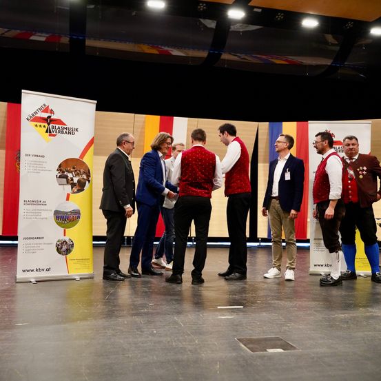 Sieben Personen stehen auf einer Bühne, gekleidet in formeller und traditioneller Kleidung. Sie sind wahrscheinlich in eine Diskussion oder Präsentation verwickelt. Die Bühne ist mit Bannern dekoriert, die Text und Bilder enthalten.