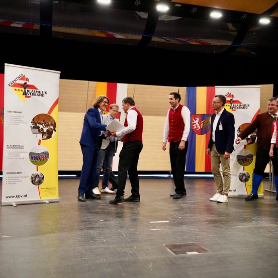 Mehrere Männer stehen auf einer Bühne, vermutlich bei einem Wettbewerb. Ein Mann in einem blauen Anzug überreicht einem Mann in einem roten Westchen ein Zertifikat. Hinter ihnen hält ein weiterer Mann ein Musikinstrument. Werbebanner befinden sich an der Seite.