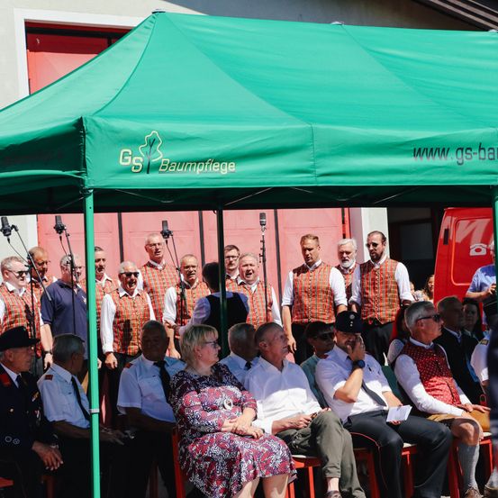 Eine Gruppe von Menschen sitzt unter einem grünen Zelt und trägt rote Westen und Krawatten. Hinter ihnen stehen mehrere Männer, die möglicherweise singen. Mikrofone sind vor ihnen aufgestellt.