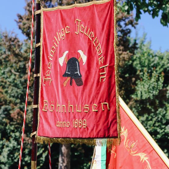 Ein rotes Banner mit goldenen Rändern und Text. Der Banner trägt 'Feuerwehr' mit gekreuzten Äxten und einem Helm. Es steht 'Bonnhusen anno 1889'. Es hängt von einem Seil.