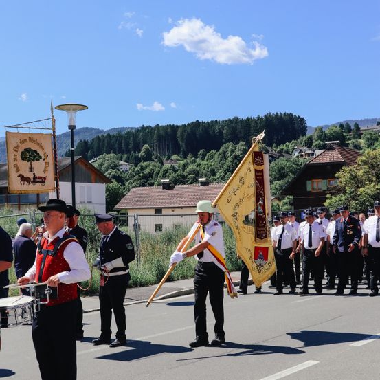 Eine Blaskapelle spielt auf der Straße. Ein Mann mit Trommel und andere sind in Uniformen gekleidet. Eine gelbe Flagge ist vorne. Gebäude und Bäume sind im Hintergrund.