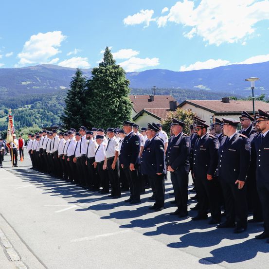 Eine Reihe uniformierter Feuerwehrleute steht an einem sonnigen Tag in Formation, mit Bergen und Häusern im Hintergrund.