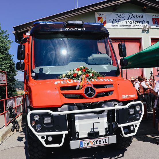Ein roter Mercedes-Benz Feuerwehrwagen, geschmückt mit Blumen, steht vor einer Feuerwehrstation. Der Wagen trägt das Wort 'FEUER' auf der Front und das Kennzeichen 'FW 266 VL'. Über dem Gebäude hängt ein Banner mit der Aufschrift '150 Jahre FCT-Paternion'.