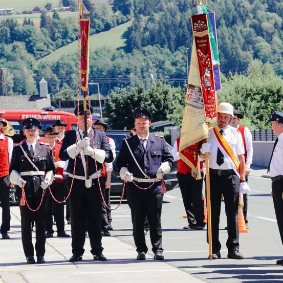 Uniformierte Personen stehen auf der Straße und halten Flaggen, wahrscheinlich während einer Parade, mit Bergen im Hintergrund.