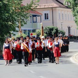 Eine Blaskapelle zieht in einem Umzug mit Blech- und Holzblasinstrumenten auf einer Stadtstraße. Eine Frau in einem roten Kleid führt den Zug an.