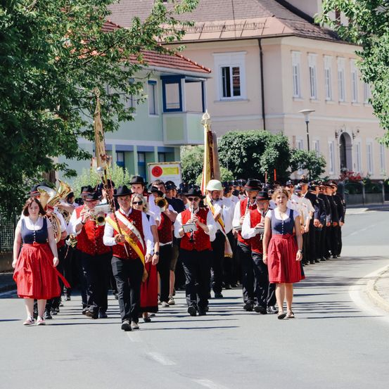 Eine Blaskapelle zieht in einem Umzug mit Blech- und Holzblasinstrumenten auf einer Stadtstraße. Eine Frau in einem roten Kleid führt den Zug an.