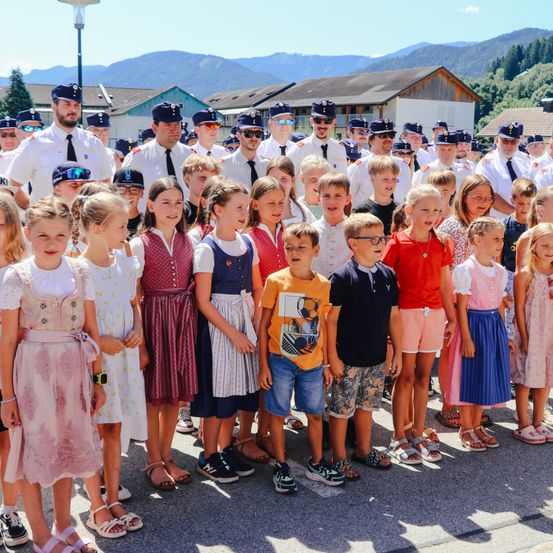 Eine Gruppe uniformierter Erwachsener und Kinder steht im Freien zusammen, einige in traditioneller Kleidung. Sie stehen in einer Reihe vor einem Gebäude mit Bergen im Hintergrund.