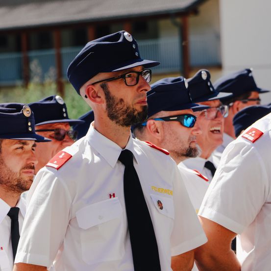 Eine Gruppe von Feuerwehrleuten in Uniform, einige mit Brillen, steht zusammen. Das Gebäude im Hintergrund hat einen Balkon und Geländer.