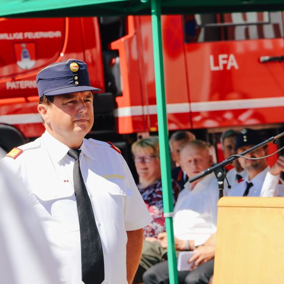 Ein Mann in Uniform steht vor einer Menge, mit einem roten Feuerwehrwagen im Hintergrund, der mit LFA beschriftet ist. Zuschauer beobachten von unter einem grünen Zelt aus, einer hält ein Mikrofon.