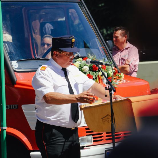Ein Feuerwehrmann spricht an einem Podest vor einem orangefarbenen Feuerwehrwagen, während ein Mann auf dem Beifahrersitz zuschaut.