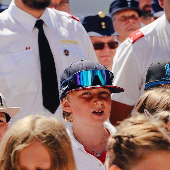 Ein junges Mädchen mit einer Kappe und einer Sonnenbrille steht unter Erwachsenen in Uniform und schaut nach oben. Einige tragen Hüte mit roten Abzeichen, während ein Mann hinter ihr einen schwarzen Krawatte trägt.
