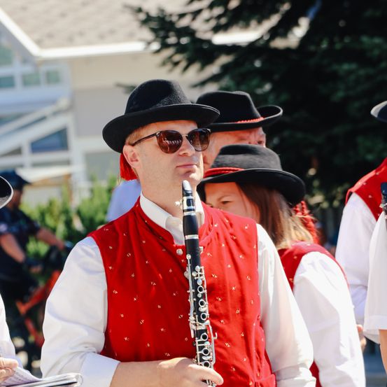 Ein Mann in einem roten Weste und schwarzem Hut spielt Klarinette bei einer Outdoor-Veranstaltung mit anderen Musikern in traditioneller Kleidung.
