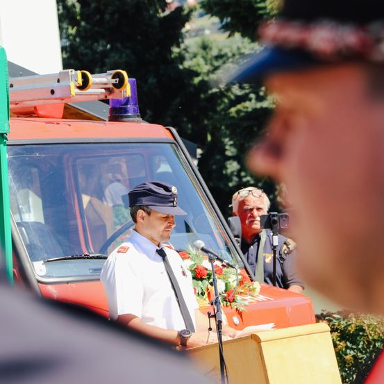 Ein Mann in Uniform steht vor einem Feuerwehrauto am Rednerpult und spricht in ein Mikrofon. Ein weiterer Mann in Uniform filmt.