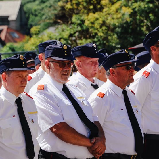Mehrere Männer in Uniform mit Mützen und Krawatten stehen in einer Reihe, möglicherweise bei einer Zeremonie. Dahinter sind Bäume und ein Haus mit einem Fenster.