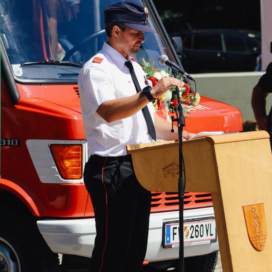 Ein Mann in Uniform steht an einem Rednerpult und spricht in ein Mikrofon. Er hält einen Blumenstrauß. Hinter ihm steht ein orangefarbenes Fahrzeug mit der Nummer 310 an der Seite.
