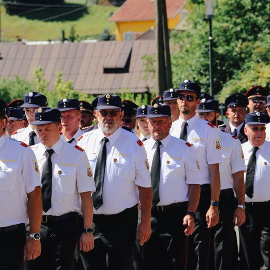 Eine Gruppe uniformierter Beamter steht in einer Reihe, nach vorne gerichtet. Sie tragen Mützen und Krawatten, und einige haben Brillen. Bäume und Gebäude sind im Hintergrund.