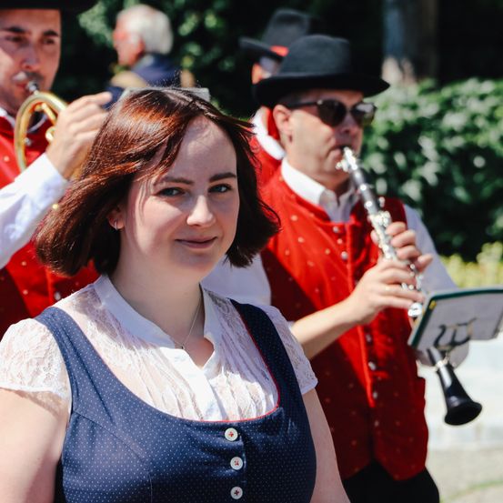 Eine Frau mit braunen Haaren lächelt und steht vor einer Band. Hinter ihr spielen zwei Männer Instrumente. Einer spielt Trompete, der andere Klarinette.