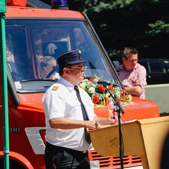 Ein Feuerwehrmann spricht an einem Podium vor einem Feuerwehrauto. Ein Mann beobachtet ihn hinter dem Auto.