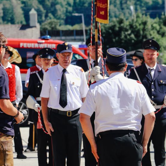Uniformierte Beamte, einschließlich Feuerwehrleute, stehen in einer Reihe mit Fahnen und Instrumenten, wahrscheinlich bei einer Zeremonie. Im Hintergrund befindet sich ein rotes Fahrzeug und ein klarer blauer Himmel.