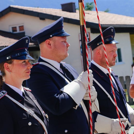 Drei Personen in Uniform, zwei Männer und eine Frau, stehen vor einem Haus. Sie halten einen Flaggenmast mit roten und weißen Seilen.