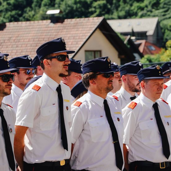Eine Gruppe uniformierter Männer steht in einer Reihe, einige mit Sonnenbrillen, möglicherweise bei einer zeremoniellen Veranstaltung. Sie tragen Mützen und schwarze Krawatten.