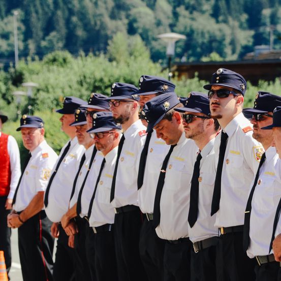 Eine Gruppe uniformierter Männer steht in einer Reihe im Freien, möglicherweise Polizeibeamte. Sie tragen Hüte, Sonnenbrillen und Krawatten. Der Hintergrund zeigt grüne Bäume und ein Gebäude.