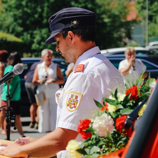 Ein Mann in Uniform spricht in ein Mikrofon, mit Menschen und Autos im Hintergrund. Er trägt eine Mütze und einen Abzeichen.