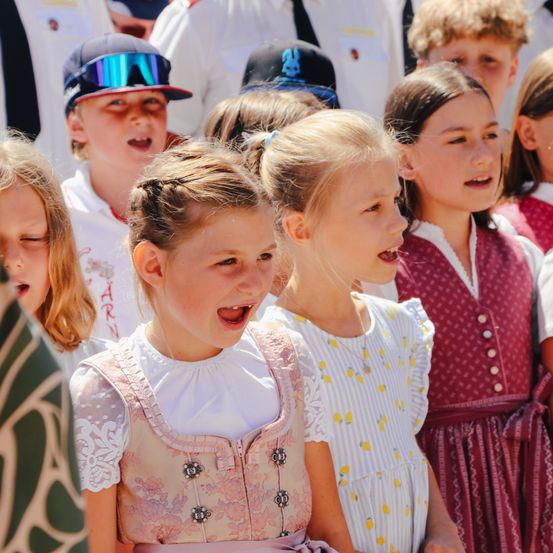 Eine Gruppe von Kindern in traditioneller Kleidung, zwei davon lächelnd, scheinen in einem Chor zu singen.