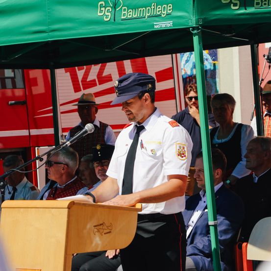 Ein Mann in Uniform spricht an einem Rednerpult unter einem Zelt, mit einem Mikrofon und einem Publikum hinter ihm. Die Menschen schauen aufmerksam zu, und ein Feuerwehrauto ist im Hintergrund sichtbar.