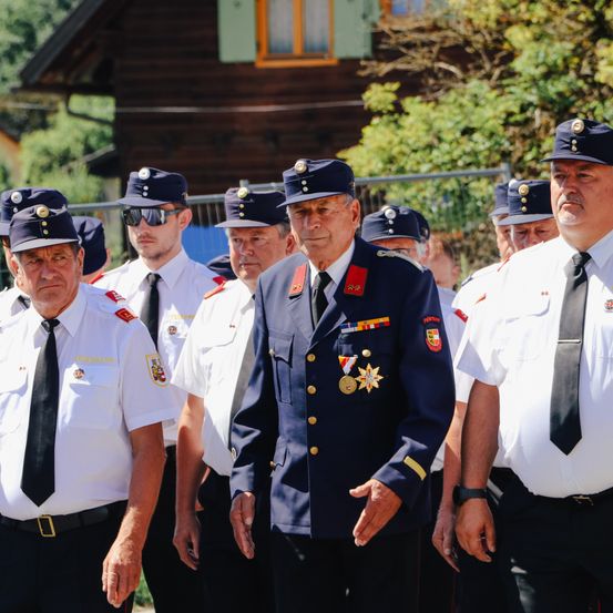 Eine Gruppe uniformierter Männer in Mützen und Krawatten steht in einer Reihe. Ein Mann in blauer Uniform führt an. Sie sind draußen in der Nähe eines Holzhauses und eines Zauns.
