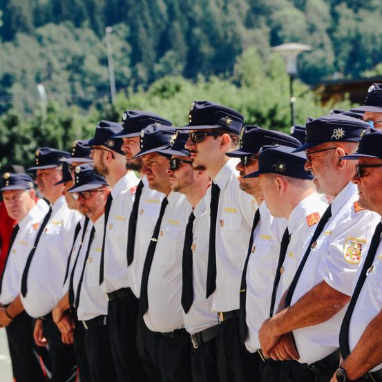 Eine Reihe von Männern in Uniform, möglicherweise Polizeibeamte, stehen in Formation im Freien, mit Bäumen und einem Gebäude im Hintergrund.