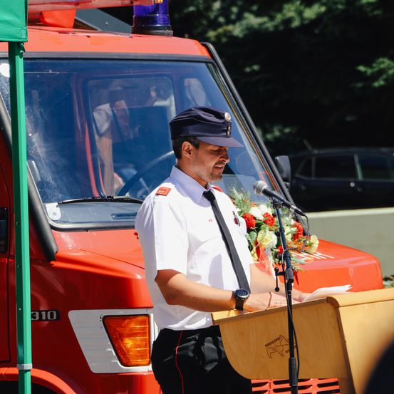 Ein Mann in Uniform spricht in ein Mikrofon vor einem Feuerwehrauto. Ein Blumenstrauß liegt auf dem Podium.