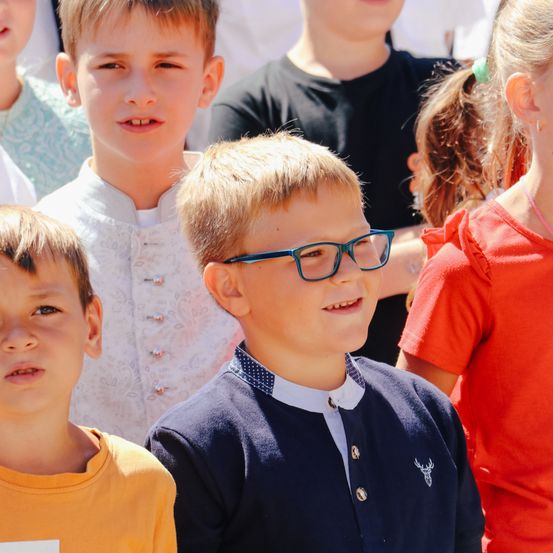 Eine Gruppe von Kindern steht zusammen; der Junge mit blauen Brillen lächelt, während andere zusehen.