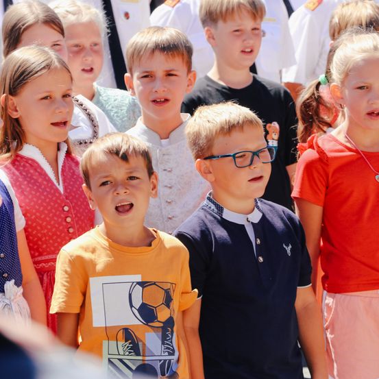 Eine Gruppe von Kindern steht in einer Reihe und singt gemeinsam. Sie tragen verschiedenfarbige Shirts und einige haben eine Brille. Im Hintergrund stehen weitere Kinder.