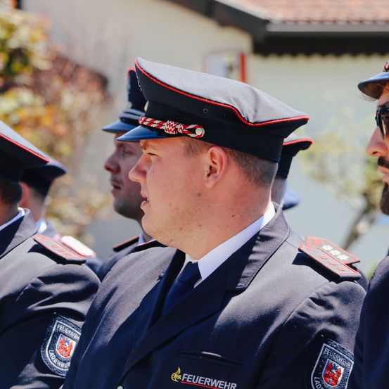 Fünf Feuerwehrleute in Uniform, mit Mützen, stehen in einer Reihe, mit einem verschwommenen Hintergrund. Der Mann in der Mitte trägt einen Krawatte.