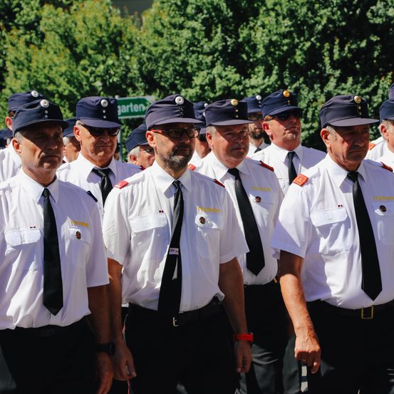 Eine Gruppe uniformierter Männer mit schwarzen Krawatten und Hüten steht in einer Reihe im Freien. Sie scheinen Sicherheitspersonal oder Beamte zu sein.