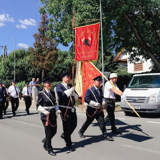 Fünf uniformierte Personen marschieren mit Fahnen die Straße hinunter. Hinter ihnen ist ein weißer Van geparkt. Menschen beobachten von der Straßenseite aus. Bäume und ein Gebäude im Hintergrund.