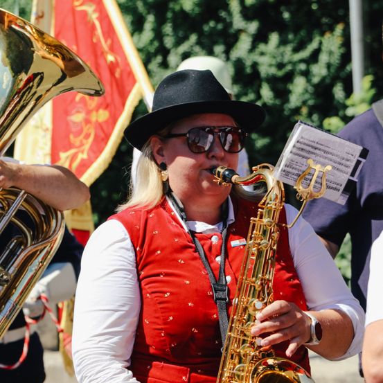 Eine Frau in einer roten Weste spielt Saxophon, mit einem Tubaspieler dahinter und einer Fahne im Hintergrund.