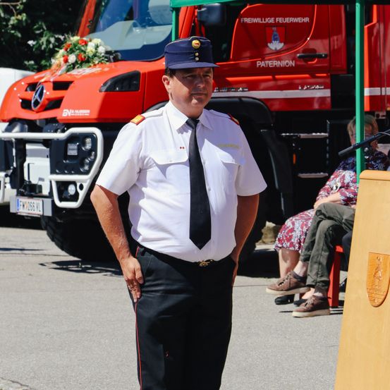 Ein Feuerwehrmann in weißer Uniform steht vor einem roten Feuerwehrauto mit einem Blumenstrauß auf der Motorhaube. Hinter ihm sitzen zwei Personen, eine mit einem Mikrofon.