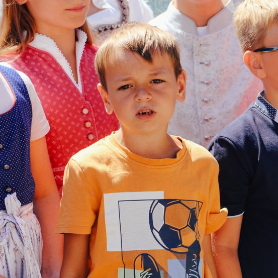 Ein junger Junge mit einem orangefarbenen Shirt mit Fußball-Motiv steht unter anderen, die traditionelle Kleidung tragen.