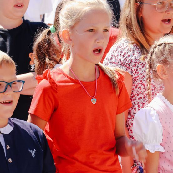 Eine Gruppe von Kindern, darunter ein Mädchen in Rot, singt gemeinsam in einer Menschenmenge. Ein Mädchen mit geflochtenen Haaren und ein weiteres Mädchen in Pink sind ebenfalls anwesend.
