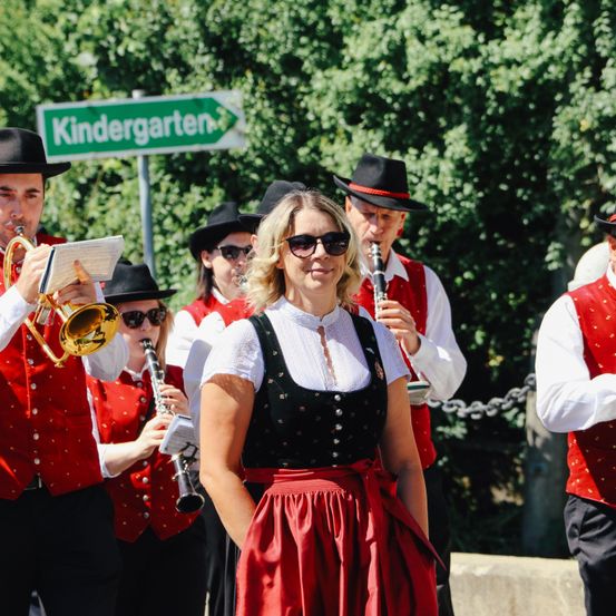 Eine Gruppe von Musikern in traditioneller deutscher Kleidung tritt draußen in der Nähe eines Schildes auf, das Kindergarten steht.