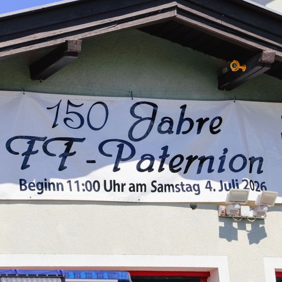 Ein Banner am Gebäude zeigt '150 Jahre FF-Paterion'. Darunter steht 'Beginn 11:00 Uhr am Samstag 4. Juli 2026'. Zwei Außenleuchten sind an der Wand befestigt.