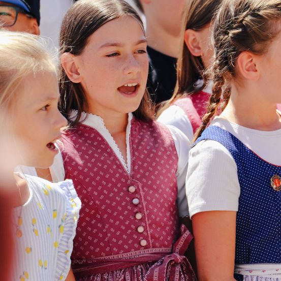 Drei junge Mädchen in traditioneller Kleidung singen gemeinsam. Das Mädchen in der Mitte trägt eine burgunderfarbene Weste mit Knöpfen und einem weißen Kragen.