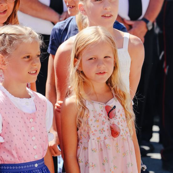 Zwei junge Mädchen mit blonden Haaren stehen eng beieinander. Sie tragen hellrosa Kleider. Das Mädchen auf der linken Seite trägt ein Kleid mit Knöpfen. Das Mädchen auf der rechten Seite hat eine Sonnenbrille, die an ihrem Kleid hängt. Hinter ihnen stehen andere Leute.