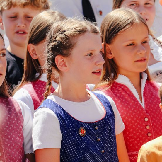 Eine Gruppe von Kindern in übereinstimmenden Uniformen, einige mit geflochtenen Haaren, stehen zusammen und schauen in die gleiche Richtung. Das Mädchen in der Mitte trägt ein blaues und weißes Kleid mit Punkten und einer Plakette.
