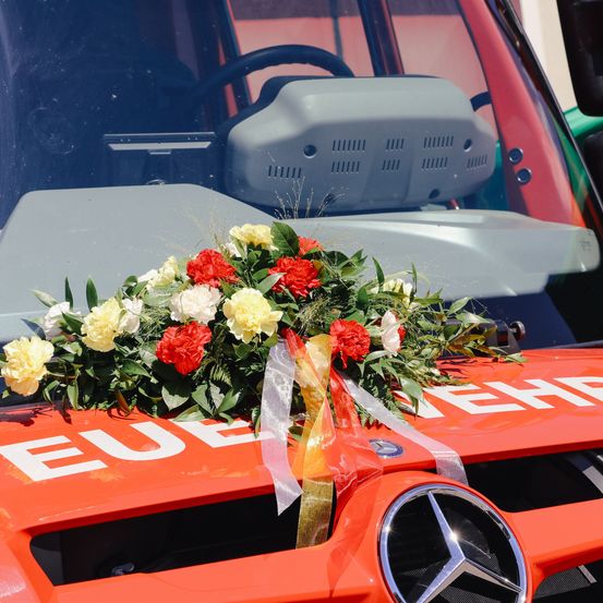 Ein orangefarbener Mercedes-Fahrzeug, möglicherweise ein Feuerwehrwagen, geschmückt mit einem Blumenstrauß in Rot, Weiß und Gelb. Das Wort 'EUE' ist prominent auf dem Fahrzeug zu sehen.