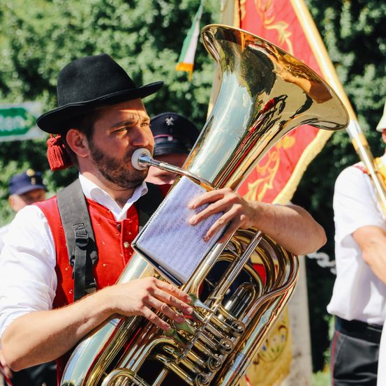 Ein Mann in traditioneller Kleidung spielt in einer Parade eine Tuba, mit einer Flagge und anderen Musikern im Hintergrund.