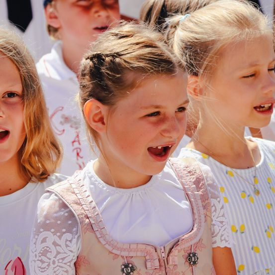 Drei Mädchen stehen eng beieinander und singen. Das Mädchen in der Mitte hat den Mund offen und lächelt. Die anderen lächeln ebenfalls.