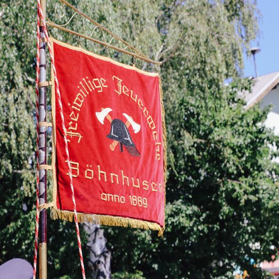 Ein rotes Banner mit goldenem Text und einem gekreuzten Axt-Design, beschriftet mit 'Freiwillige Feuerwehr Böhnhusen anno 1889'. Es ist an einem Mast befestigt vor einem Hintergrund aus grünen Bäumen und einem weißen Gebäude.